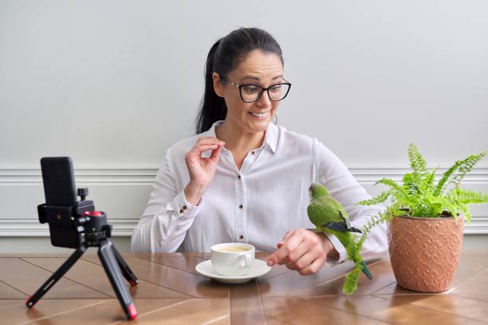 house sitter sitting in front of a camera on a tripod wearing glasses and a white shirt looking at green bird perched on their hand next to a coffee and a pot plant